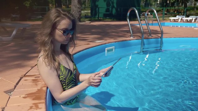 blonde woman is viewing pictures in tablet, resting in water of swimming pool in sunny weather