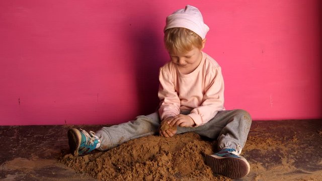 Little blond boy in pink sweater, hat and blue boots enthusiastically plays with sand. Beautiful chubby baby hands close-up. Kid comes up with entertainment and actively masters necessary skills