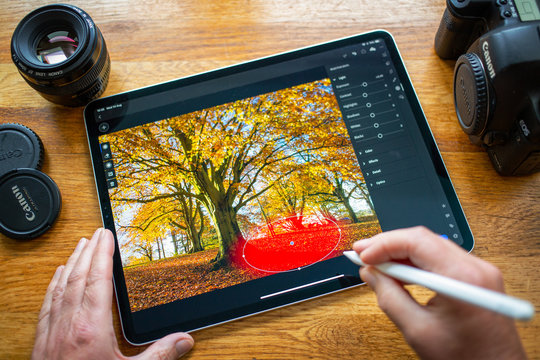 BATH, UNITED KINGDOM - AUGUST 14, 2019 : Close up of someone using an iPad Pro running the Adobe Lightroom App to make selective adjustment to a photograph of colourful trees.