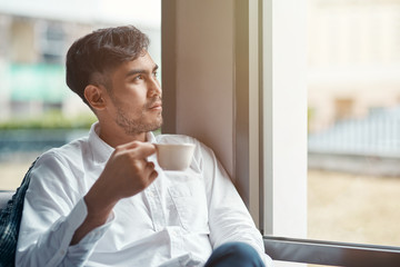 Portrait of asian male worker in white shirt sitting in the modern office, looking at the window and drinking coffee during lunch break. Horizontal shot