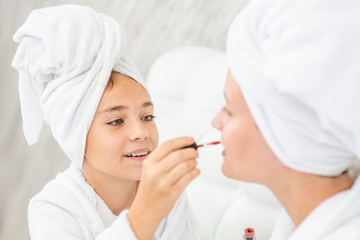 Happy family at home. Mother and daughter are doing make up and having fun sitting on the bed at home. Mom and child girl are in bathrobes and with towels on their heads