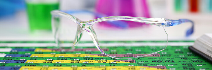 Protective surgical or lab glasses lying on periodic table closeup and sample bottle with poison fluid in background