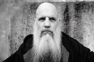 Mature bald man with long gray beard against concrete wall outdoors