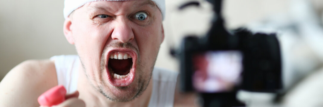 Male vlogger holds dumbbells in his hands and shouts at the camera on his subscriber, motivating them to action. Tutoring grade on an individual program online fraud concept aganist home background.