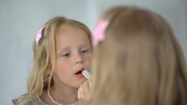 Little funny girl applying red lipstick, doing make up looking at mirror and smiling. child at mirror with lipstick, playing with mothers makeup, little fashionista doing make-up painting her lips 4 K