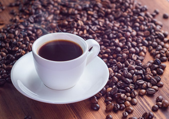 Heap of natural coffee beans and white cup full of black espresso on brown table, close up view. Texture of brown desk. Selective soft focus. Blurred background