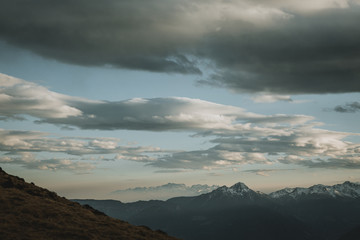 Panorama of snowy peaks of the mountains with beautiful cloud sky above them during orange sunset with haze in the valley with yellow blurry meadow in front