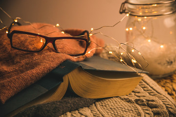 Cozy warmly lit reading area or nook for winter and fall weather with led string lights, books, glasses, wicker table and wool blanket or throw - obrazy, fototapety, plakaty