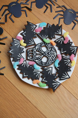 Halloween cookies in shape of skull and bat with colorful worm candies on a plate on wooden background. Halloween sweet food