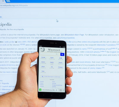 Adygea, Russia - January 5, 2018: American Public Multilingual Universal Internet Encyclopedia With Free Content Wikipedia On The Screen Of The Chinese Phone Xiaomi In The Male Hand And On Monitor