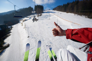 Winter mountain landscape. Close-up of skier hand with thumb-up gesture and two pairs of skis on background of rope way on sunny winter day. Sports and recreation, active lifestyle concept.