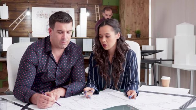 Businesswoman Talking With Her Contructor Engineer With Blueprints On The Office. Constructor Engineer Drawing On Blueprints.