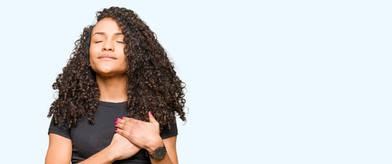 Young beautiful woman with curly hair smiling with hands on chest with closed eyes and grateful...