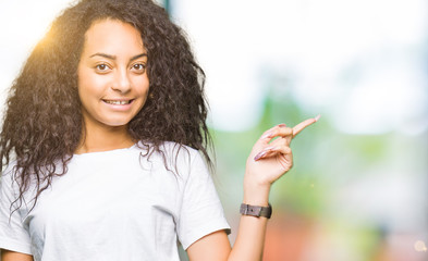 Young beautiful girl with curly hair wearing casual white t-shirt with a big smile on face, pointing with hand and finger to the side looking at the camera.