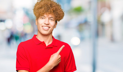 Young handsome man with afro hair wearing red t-shirt cheerful with a smile of face pointing with hand and finger up to the side with happy and natural expression on face