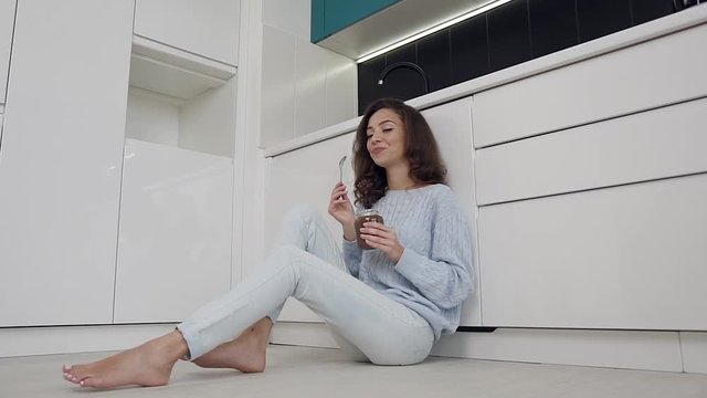 Adorable modern smiling woman in stylish clothes sitting on the kitchen floor and eating chocolate cream from jar