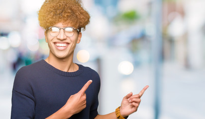 Young handsome man with afro wearing glasses smiling and looking at the camera pointing with two hands and fingers to the side.