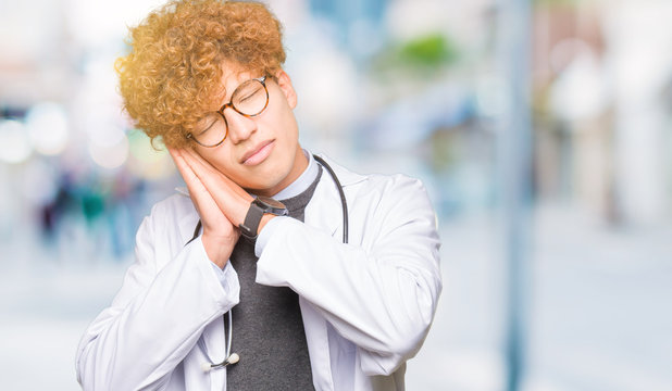 Young handsome doctor man wearing medical coat sleeping tired dreaming and posing with hands together while smiling with closed eyes.