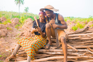 excited african couple doing internet banking on their farmland. male and female farmers using smartphone   - obrazy, fototapety, plakaty