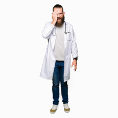 Young blond doctor man with beard wearing medical coat covering one eye with hand with confident smile on face and surprise emotion.