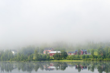 Fototapeta na wymiar Farm at a mirrored lake and fog in a forest landscape