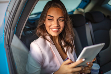 Cheerful young woman reading messages in Tablet PC while sitting in a taxi. Cute young woman traveling. Focused businesswoman using her tablet PC in her car.