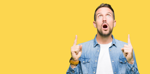 Handsome man with blue eyes and beard wearing denim jacket amazed and surprised looking up and pointing with fingers and raised arms.