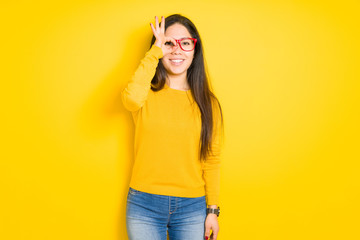 Beautiful brunette woman wearing red glasses over yellow isolated background doing ok gesture with hand smiling, eye looking through fingers with happy face.