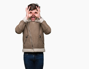 Young handsome man wearing winter coat over isolated background doing ok gesture like binoculars sticking tongue out, eyes looking through fingers. Crazy expression.
