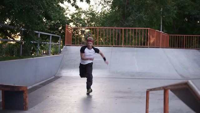 Sportive man practicing parkour in the open skate park - sliding from the ramp, crossing the barries amd making somersault in slow motion. Summer day, green trees around