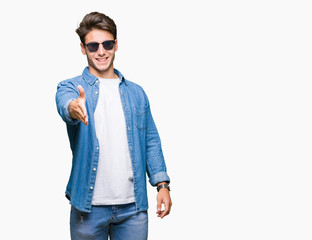 Fototapeta premium Young handsome man wearing sunglasses over isolated background smiling friendly offering handshake as greeting and welcoming. Successful business.