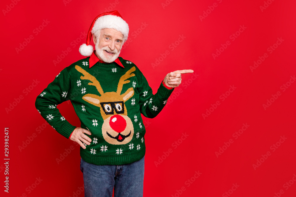 Wall mural Photo of senior man indicating finger to empty space advising shocking low december prices wear x-mas ugly ornament sweater santa cap isolated red background