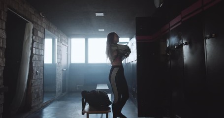 Beautiful young happy Caucasian woman changing clothes after gym exercise in dark locker room, life motivation concept.