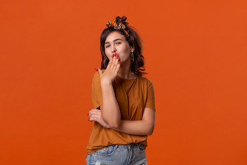 Pretty brunette woman in a t-shirt and beautiful headband sends air kisses standing isolated over orange background. Concept of love