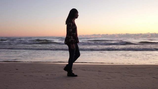 Slow motion clip of an African American woman walking across frame along a sandy beach shoreline as waves roll in at sunrise with pink hues to the sky and clouds at the horizon beyond.