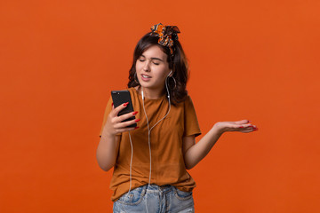 Pretty brunette woman in a t-shirt and beautiful headband is displeased video chatting on the smartphone isolated over orange background. Daily communication