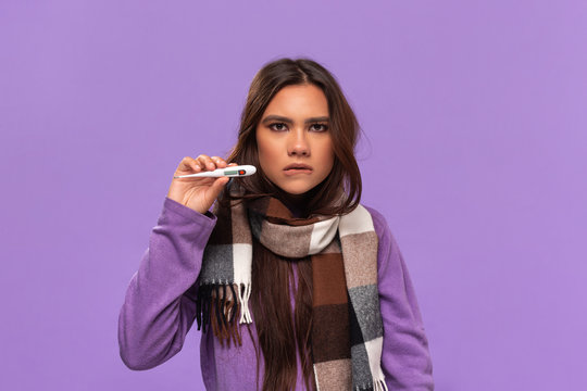 Pretty african-american girl in a sweater and cosy checked scarf checking thermometer standing isolated over purple background. Catching cold in winter.