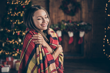 Profile side photo of positive cheerful brunette hair girl covered by checkered blanket hug herself feel warmth soft comfort in house with christmas decor lights indoors