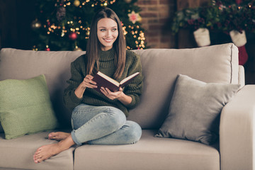 Full body photo of positive cheerful girl sit cozy divan cross her legs read christmas story imagine enjoy newyear vacation party celebration in house full of x-mas ornament indoors