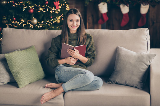 Full size photo of positive cheerful brown hair girl sit on divan hold book enjoy christmas celebration vacation wear knitted sweater denim jeans in house full of noel decorations tinsels indoors