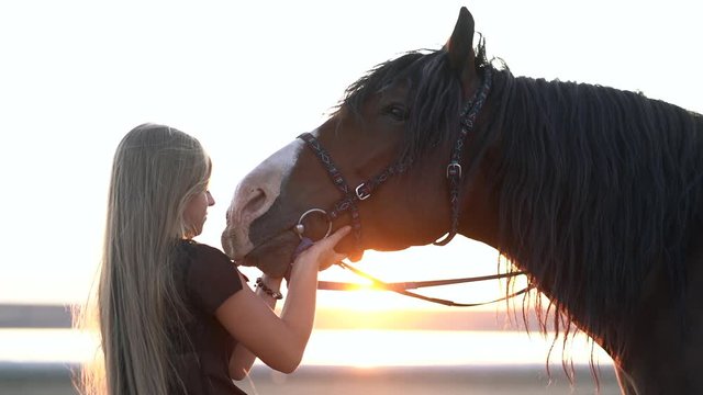 Young blonde girl with long hair stroking and kissing horse. Slow motion. Beautiful young woman with dark stallion on nature. Love and friendship concept.