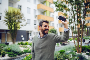 Smiling handsome fashionable Caucasian man standing in park, holding smart phone and waving.