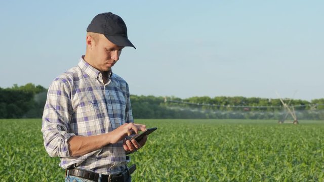Young male farmer uses a mobile app in the smartphone in the field against the background of a working irrigation system in the distance, soft focus