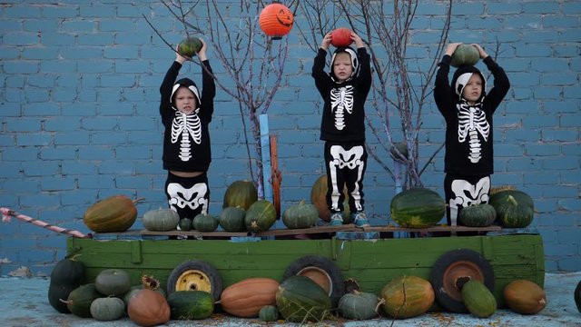 Children in carnival costumes of skeletons for Halloween are sitting in boat with pumpkins. Brothers gathered good crop of pumpkins in garden and put them on ship. holiday suite in village