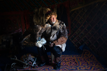 Portrait of an old kazakh eagle hunter with his majestic golden eagle indoors in dark traditional...