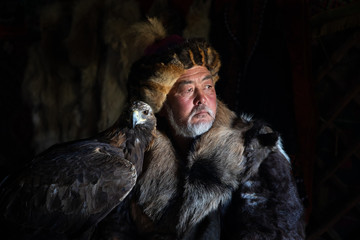 Portrait of an old kazakh eagle hunter with his majestic golden eagle indoors in dark traditional...