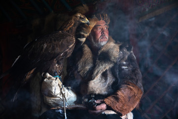 Portrait of an old kazakh eagle hunter with his majestic golden eagle indoors in dark traditional...