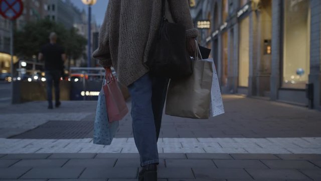Low-angle handheld shot of a young woman walking with colorful shopping bags in the city.