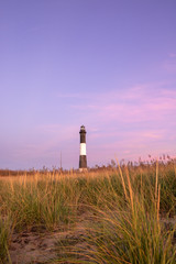 Fototapeta na wymiar Soft pink pastel colors in the sky behind a lighthouse with warm light still on the beachgrass. Fire Island Lighthouse, New York