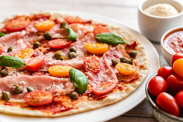 Italian pizza with prosciutto ham, capers, cherry tomatoes, parmesan cheese and decorated with basil leaves.